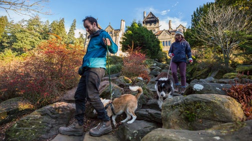 Two visitors walk two dogs on leads down the Rock Garden with the house in the background on a sunny autumn day at Cragside, Northumberland